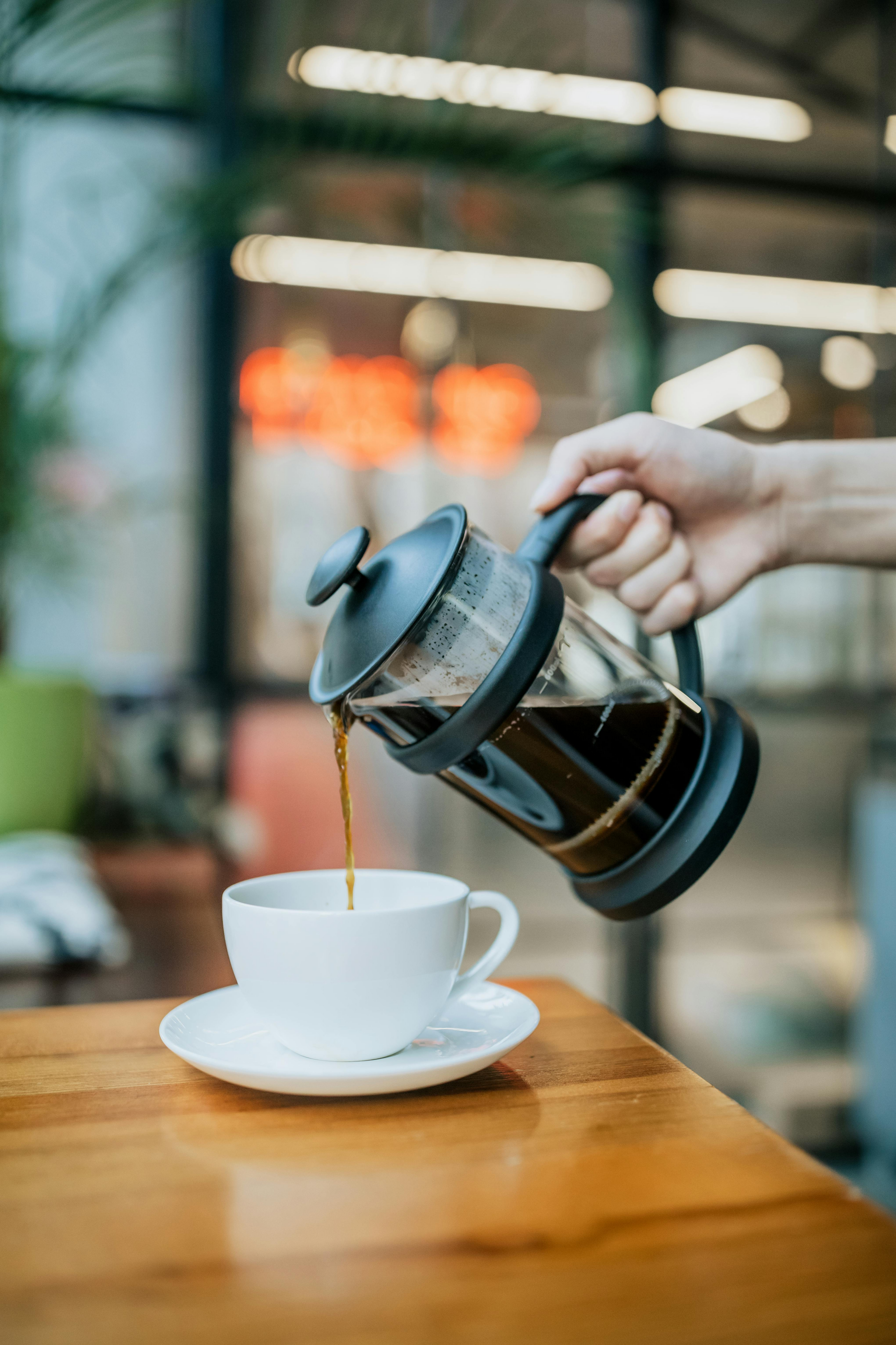 Person pouring coffee from a French press into a white cup on a wooden table.