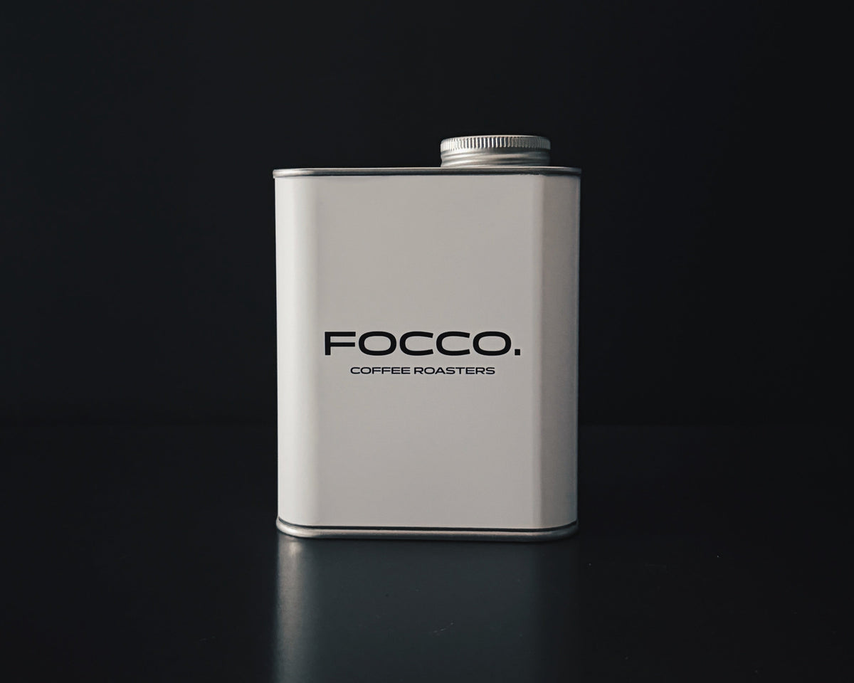 A matte white rectangular metal tin with a silver screw cap, standing upright on a dark reflective surface. The front of the tin is printed with the brand name 'FOCCO.