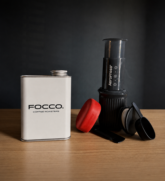 A Focco coffee roasting kit arranged on a wooden tabletop against a dark studio background. The set includes a white metal canister labeled FOCO.