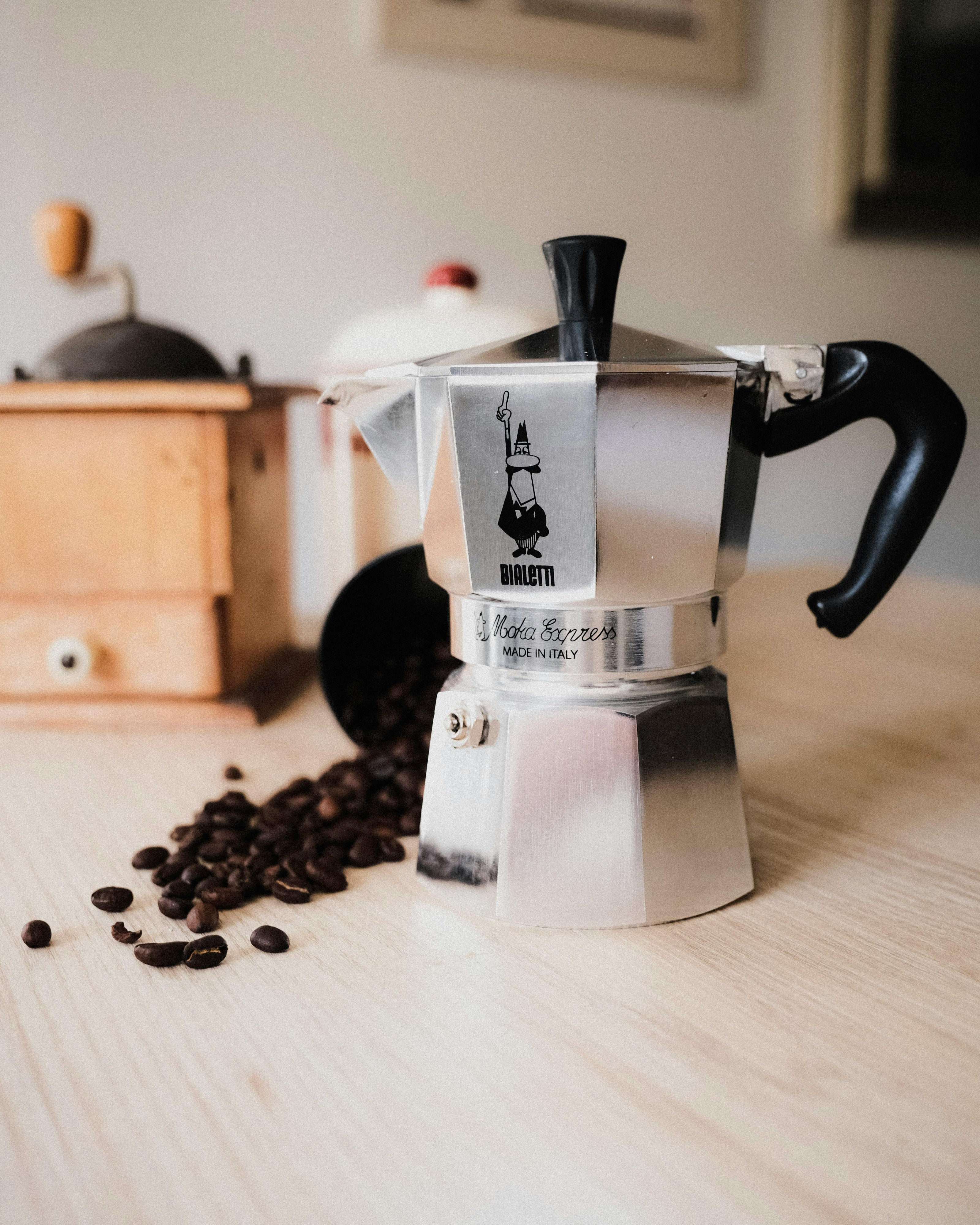 Silver Moka pot with black handle on a wooden surface with coffee beans and a wooden grinder in the background.
